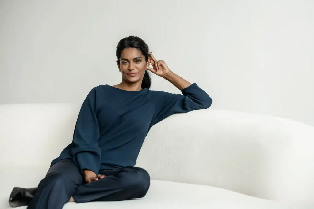 Eine Frau sitzt entspannt auf einem weißen Sofa und trägt ein dunkelblaues Outfit, während sie mit ruhigem Blick in die Kamera schaut.
